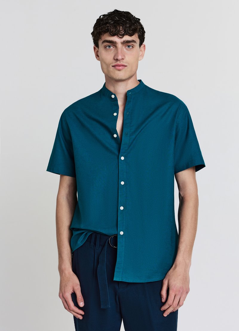 Chemise bleu sarcelle à manches courtes avec col mao, boutons sur le devant et texture lisse. Associée à un pantalon foncé avec une ceinture nouée.