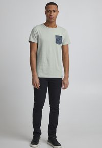 Tricou din bumbac verde deschis cu un buzunar modelat în albastru închis, asociat cu blugi negri slim-fit și adidași negri. Design simplu și casual.