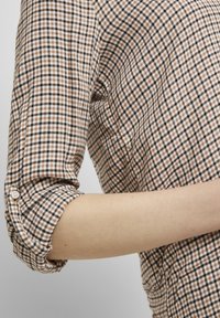 Chemise à carreaux marron et blanc avec les manches roulées, dotée d'une fermeture à boutons et d'un tissu texturé. Coupe cintrée, design subtil.