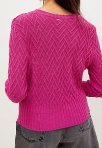 Gebreide trui in helderroze met een gestructureerd herringbone-patroon, geribbelde zoom en zilveren accentknopdetail op de schouder.