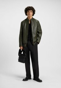Giacca di pelle verde oliva, chiusura con zip, colletto rotondo, indossata su una maglietta nera. Pantaloni neri e una piccola borsa nera con dettagli del logo.