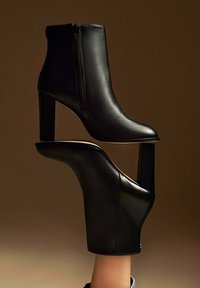 Paire de bottines en cuir noir élégantes à talons carrés, l'une portée au pied et l'autre posée au-dessus, sur un fond marron.