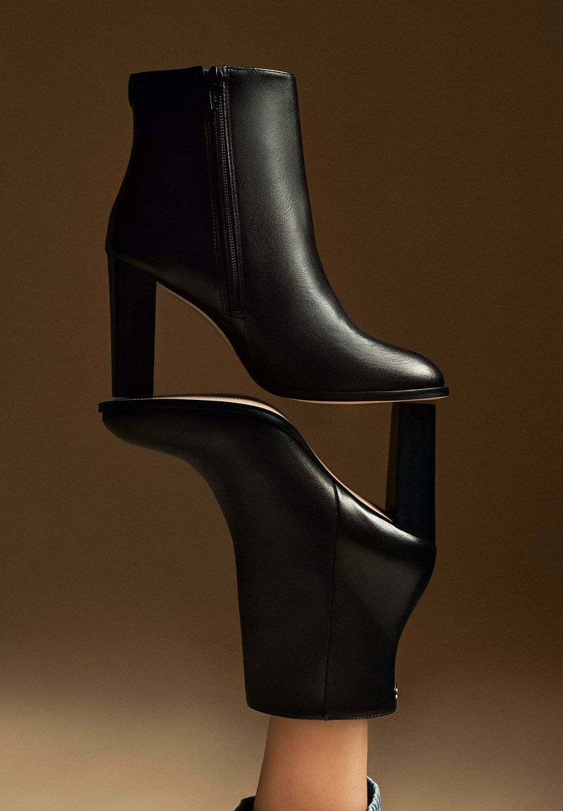 Paire de bottines en cuir noir élégantes à talons carrés, l'une portée au pied et l'autre posée au-dessus, sur un fond marron.