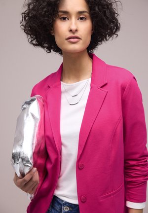 Vrouw met krullend haar draagt een felroze blazer en een wit overhemd, houdend een glanzende zilveren clutch vast tegen een lichte achtergrond.