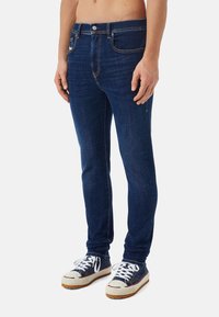 Jeans de mezclilla azul oscuro con corte recto, diseño de cinco bolsillos y costuras contrastantes. Combinados con zapatillas blancas y azul marino.