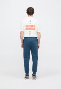 Λευκό βαμβακερό μπλουζάκι με έντονο πορτοκαλί κείμενο "BROKEN RECORDS" στην πλάτη. Φορεμένο με μπλε σκούρο αθλητικό παντελόνι και γκρι αθλητικά παπούτσια.