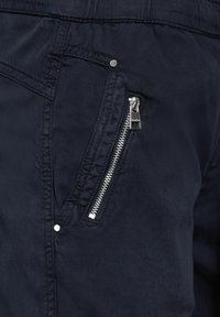 Navy lehké kalhoty s boční kapsou na zip se stříbrnými doplňky a plochým zadním pasem. Materiál má hladkou, mírně lesklou texturu.