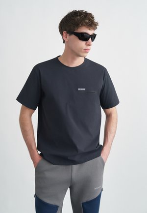 Young man wearing black sunglasses, black Columbia t-shirt with chest pocket, and gray Columbia pants with navy knee patches, hands in pockets.