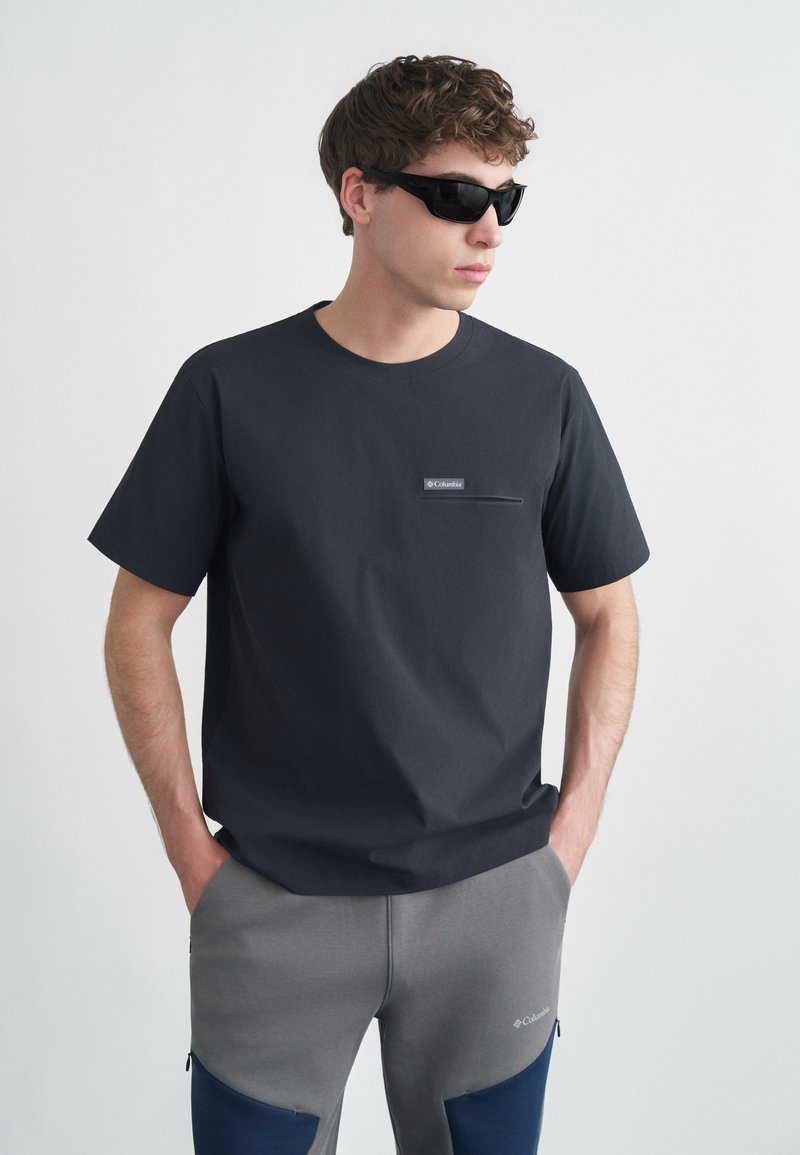 Νεαρός άνδρας που φοράει μαύρα γυαλιά ηλίου, μαύρο μπλουζάκι Columbia με τσέπη στο στήθος και γκρι παντελόνι Columbia με μπλε επιθέματα στα γόνατα, με τα χέρια στις τσέπες.