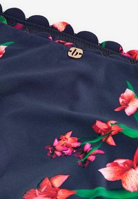 Tumši zils audums ar ziedu rakstu rozā un sarkanā krāsā ar zaļiem akcentiem. Aprīkots ar izliektu apmali un zelta toņa dekoratīvu pogu.