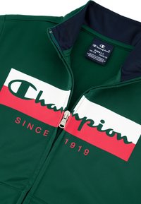 Chaqueta verde con cremallera y cuello azul marino. Logo de Champion en blanco y rojo en la parte delantera con "DESDE 1919" en letras rojas.