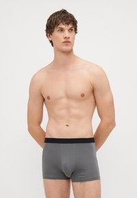 Graue Boxershorts mit schwarzem Bund, enganliegendem Design, glatter Textur und ohne sichtbare Muster. Modell hat einen nackten Oberkörper.