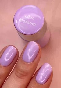 Fliederfarbener Nagellack mit glänzender Oberfläche und runder Kappe, beschriftet mit "Fliederblüte". Die Fingernägel zeigen einen einheitlichen Lavendelton.