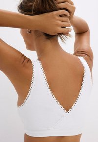Donna con capelli corti che indossa una maglietta senza maniche bianca con bordi scallop, mani appoggiate dietro la testa, vista da dietro.