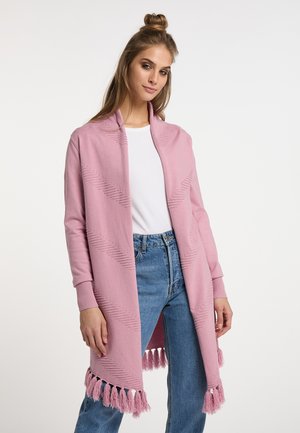 Ung kvinde iført en lyserød strikket cardigan med frynser, hvid t-shirt og blå jeans, står mod en enkel lys baggrund.