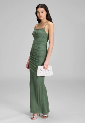 Mujer con un vestido maxi sin mangas verde oliva fruncido, sosteniendo un clutch blanco, con tacones plateados y pendientes de aro, de pie sobre un fondo gris.