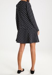 Femme debout, portant une robe noire à manches longues à pois blancs et des chaussures plates noires, photographiée de dos sur un fond blanc.