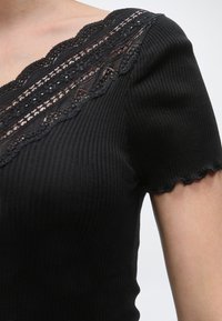 Μαύρο ριμπ κοντομάνικο τοπ με κυματιστά τελειώματα και λεπτομέρεια δαντέλας σε σχήμα V στο ντεκολτέ, που φοριέται στο πάνω μέρος του σώματος ενός ατόμου.