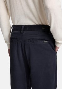 Pantaloni leggeri blu navy con una superficie liscia. Presentano una vita piatta, due tasche posteriori e un piccolo marchio sul lato destro.