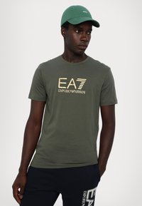 Olivenfarget bomullst-skjorte med gull- og svart logo-print. Modellen har på seg matchende mørke shorts med "EA"-merke i hvitt.