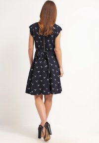 Marinblå klänning med fågeltryck, korta ärmar, figursydd midja med knytbälte och en utsvängd kjol. Bärs med svarta högklackade skor.