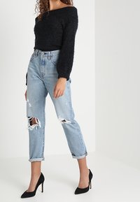 Czarny, miękki sweter z dzianiny, jasnoniebieskie jeansy z przetarciami i podwiniętymi mankietami oraz czarne szpilki. Modelka stoi na tle o neutralnym kolorze.