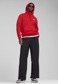 Mand iført en rød Puma hoodie, sorte vide bukser, sorte solbriller og sneakers i sort og hvid står med en ensfarvet baggrund.