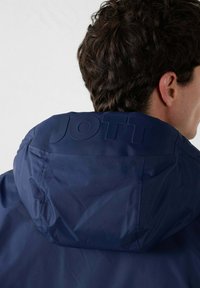 Hombre con cabello rizado oscuro que lleva una chaqueta con capucha azul marino con el logo de JOTT grabado en la parte trasera de la capucha, de espaldas.