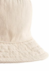 Cappello a secchiello di colore crema realizzato in tessuto di cotone testurizzato. Presenta una larga visiera cucita e un design della corona semplice e liscio.