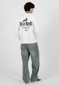 Jeune homme portant une chemise blanche à manches longues avec un logo de cheval et le texte "In a Rush", un jean bleu ample, des baskets noires et un foulard rayé autour du cou.