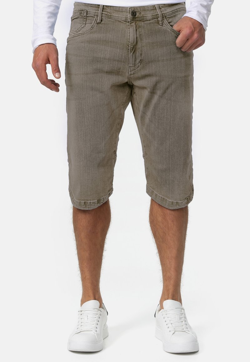 Homme portant un short en denim marron jusqu'aux genoux et des baskets blanches, debout avec une main dans la poche, sur un fond uni.
