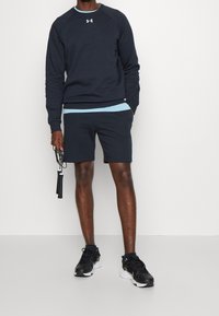 Uomo che indossa una felpa blu navy e pantaloni corti, tiene una corda per saltare nera con scarpe da ginnastica nere e bianche su uno sfondo bianco semplice.