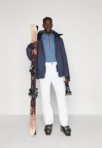 Άτομο που φοράει μπλε μπουφάν, ανοιχτό μπλε πουλόβερ, λευκά σαπούνια σκι και μαύρα μποτάκια σκι, κρατώντας σκι και γυαλιά σκι, μπροστά σε απλό φόντο.