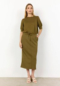 Olive zelené pletené šaty s krátkymi nariasenými rukávmi, elastickým pásom a bočnými vreckami. Hladká tkanina, dĺžka po členky, skombinované s svetlými sandálmi.