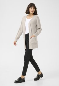 Beige cardigan met open voorkant, lange mouwen en twee voorzakken, gecombineerd met zwarte broek en glanzende zwarte schoenen. Soepele stoftextuur.
