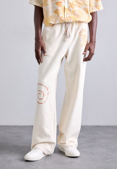 Homme portant un pantalon large texturé crème avec des imprimés en spirale orange, associé à des baskets blanches et une chemise jaune à motifs.