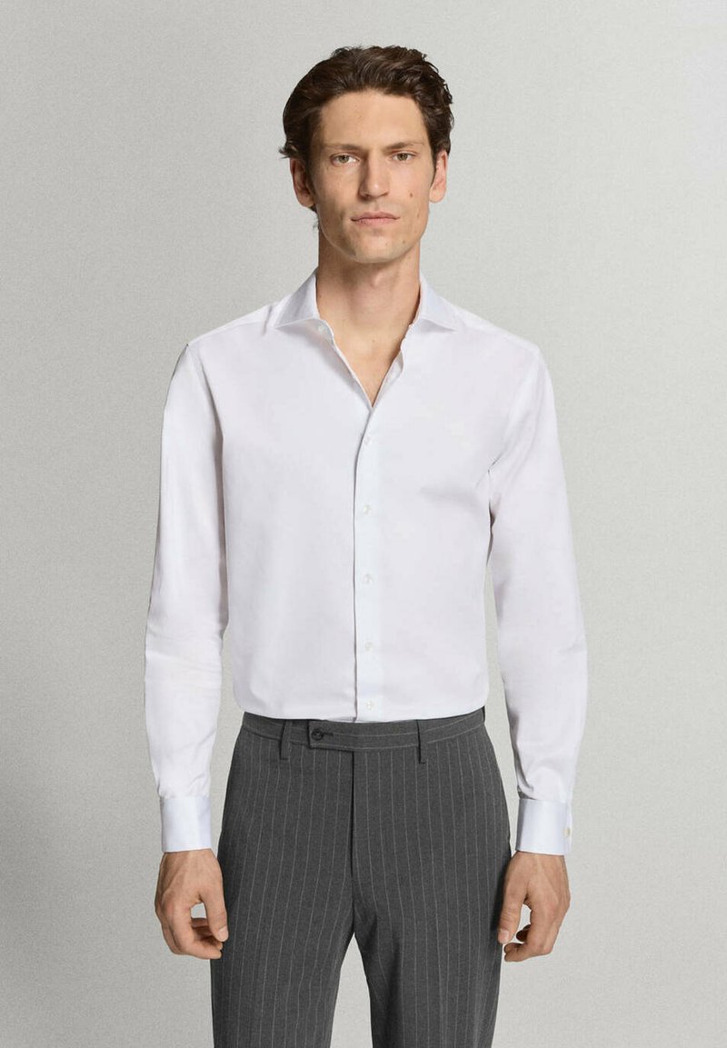 Homme portant une chemise blanche à manches longues et un pantalon gris à fines rayures, debout devant un fond uni gris clair.