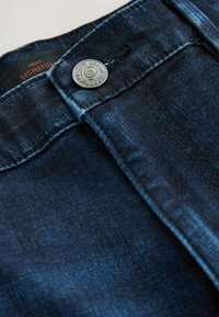 Dark blue denim jeans with a smooth texture, featuring a silver metal button, contrasting stitching, and a label inside the waistband.