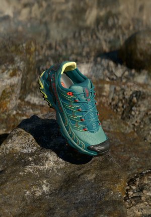Sapatilha de corrida teal com parte superior de malha texturizada, biqueira de borracha preta e detalhes em amarelo e laranja vibrantes. Leve, projetada para tração.