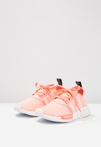 Világos rózsaszín Adidas sportcipők texturált kötött felsőrésszel, fehér talppal és passzoló fűzőkkel. Fehér csíkokkal és fekete húzófüllel rendelkezik.