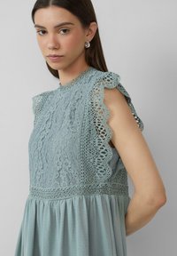 Robe en dentelle bleu clair avec des bords de manches festonnés et des motifs complexes. Elle possède un col montant et une taille froncée pour une silhouette fluide.