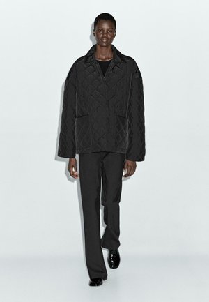 Model wearing a black quilted oversized jacket, black straight-leg trousers, and black polished shoes, standing against a plain light background.