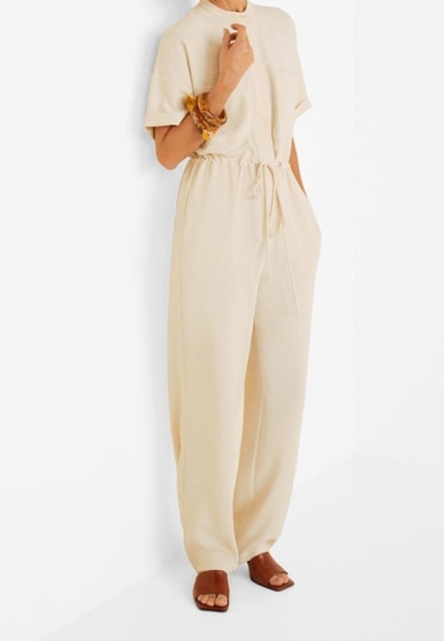 Femme portant une combinaison beige à manches courtes avec taille à cordon, des sandales marron ouvertes aux orteils, tenant sa main près de son cou.