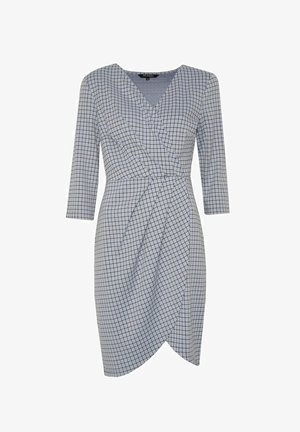 Vrouw draagt een blauw geruit wikkeljurk met driekwart mouwen, houdt een grote beige clutch vast en draagt roze sandalen met hak.