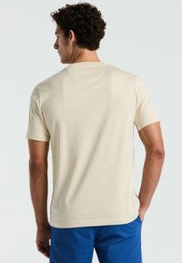 Beige t-shirt met korte mouwen, ronde hals, gladde textuur en een klassieke pasvorm. Heeft een effen achterkant en is gecombineerd met blauwe shorts.