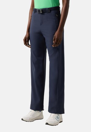 Person trägt marineblaue Hosen mit weitem Bein, schwarzen Gürtel, grünes Shirt und weiße Turnschuhe und steht vor einem einfarbigen Hintergrund.