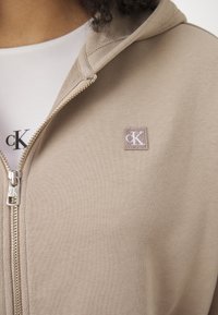 Haut à capuche beige zippé en tissu doux, avec un petit patch logo brodé et des détails en métal argenté sur la fermeture éclair.