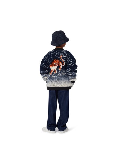 Veste sherpa bleue marine floue avec un design de tigre blanc et orange au dos, assortie à un jean foncé et un chapeau de seau foncé.