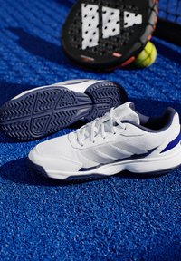 Weiße Sportschuhe mit einem strukturierten Mesh-Obermaterial, marineblauen Akzenten und einer gemusterten Gummisohle, platziert auf einer blauen synthetischen Oberfläche.