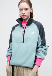 Pulover iz flisa v svetlo modri in temno sivi barvi, z zip ovratnikom. Ima roza rebraste manšete in majhen logotip na prsih.